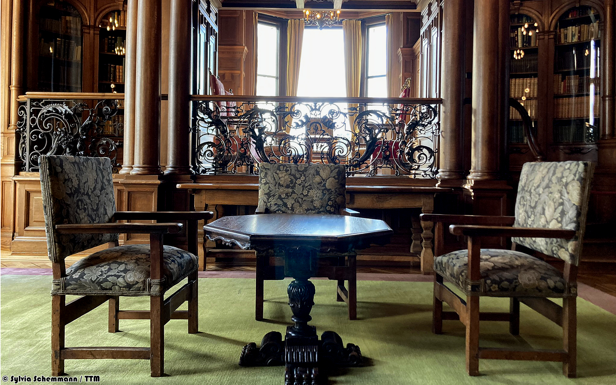 Tisch mit drei Polsterstühlen vor einem verzierten Geländer und Bibliothek im Hintergrund