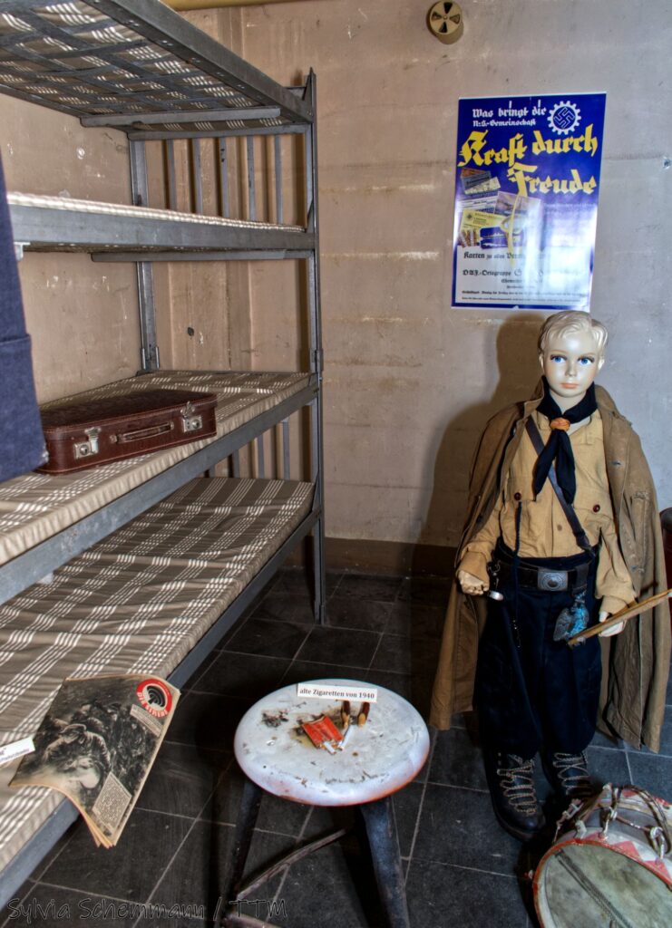 Ein kleiner Raum im Bunker mit einem vierstöckigen Etagenbett, einem Tischchen mit alten Zigaretten und einer Kinder-Schaufensterpuppe, die eine Uniform der Hitlerjugend trägt.