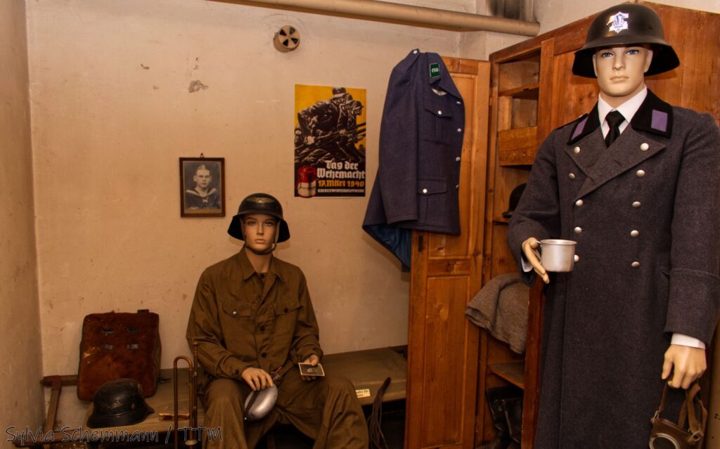 Zwei Schaufensterpuppen mit Uniformen eines Bunkerwarts und eines Luftschutzhelfers im Bunker. Beide tragen Stahlhelme.