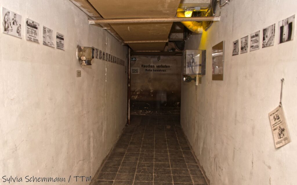 Ein Gang im Bunker mit dunkel gekacheltem Boden und weißen Betonwänden. Vor Kopf ein Schild mit der Aufschrift "Rauchen verboten".