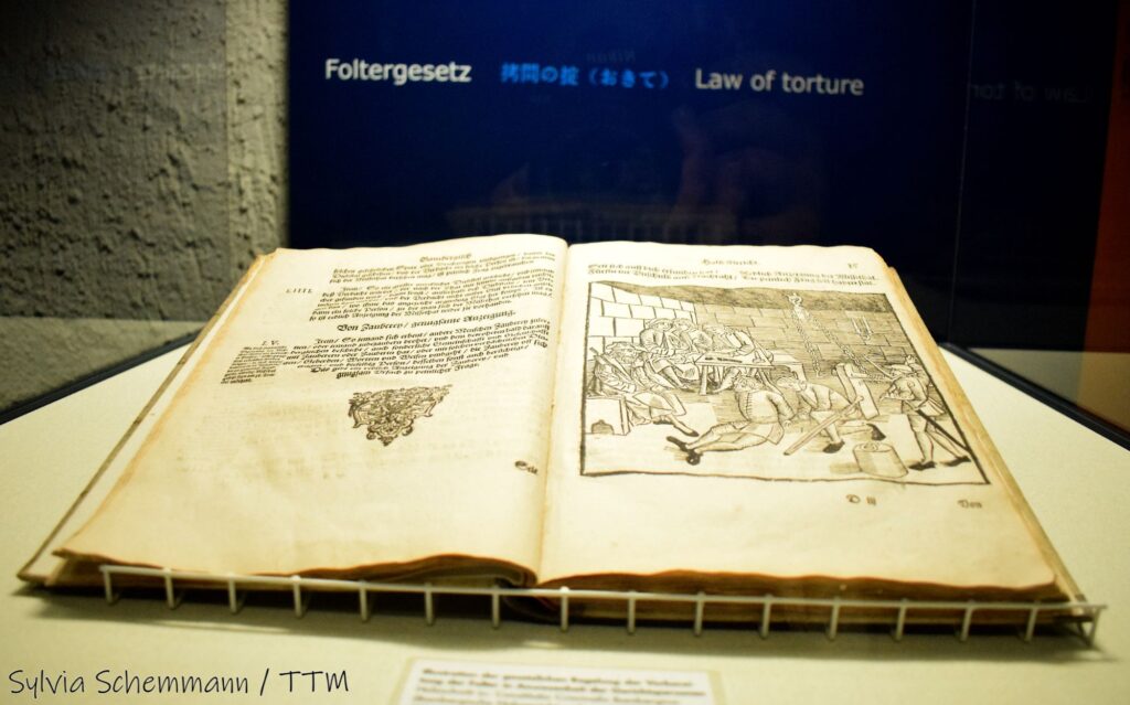 Aufgeschlagenes historisches Buch mit Schrift auf der linken Seite und einer Zeichnung, die eine Folter zeigt, auf der rechten. Dahinter eine Tafel mit der Aufschrift "Foltergesetz". Kriminalmuseum Rothenburg.