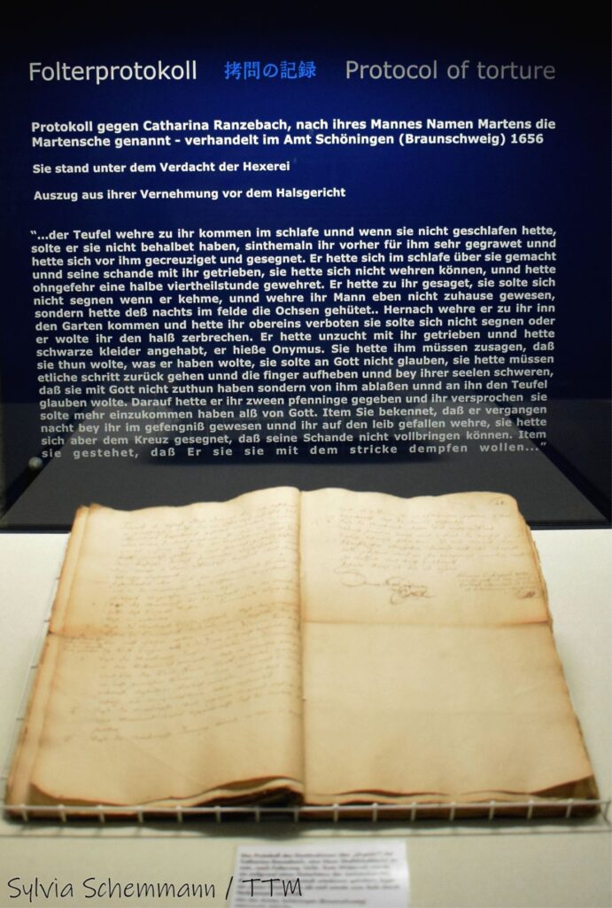 Ein aufgeschlagenes historisches Buch, dahinter eine Tafel mit der Aufschrift "Folterprotokoll" und einem Auszug aus dem ausgestellten Protokoll.