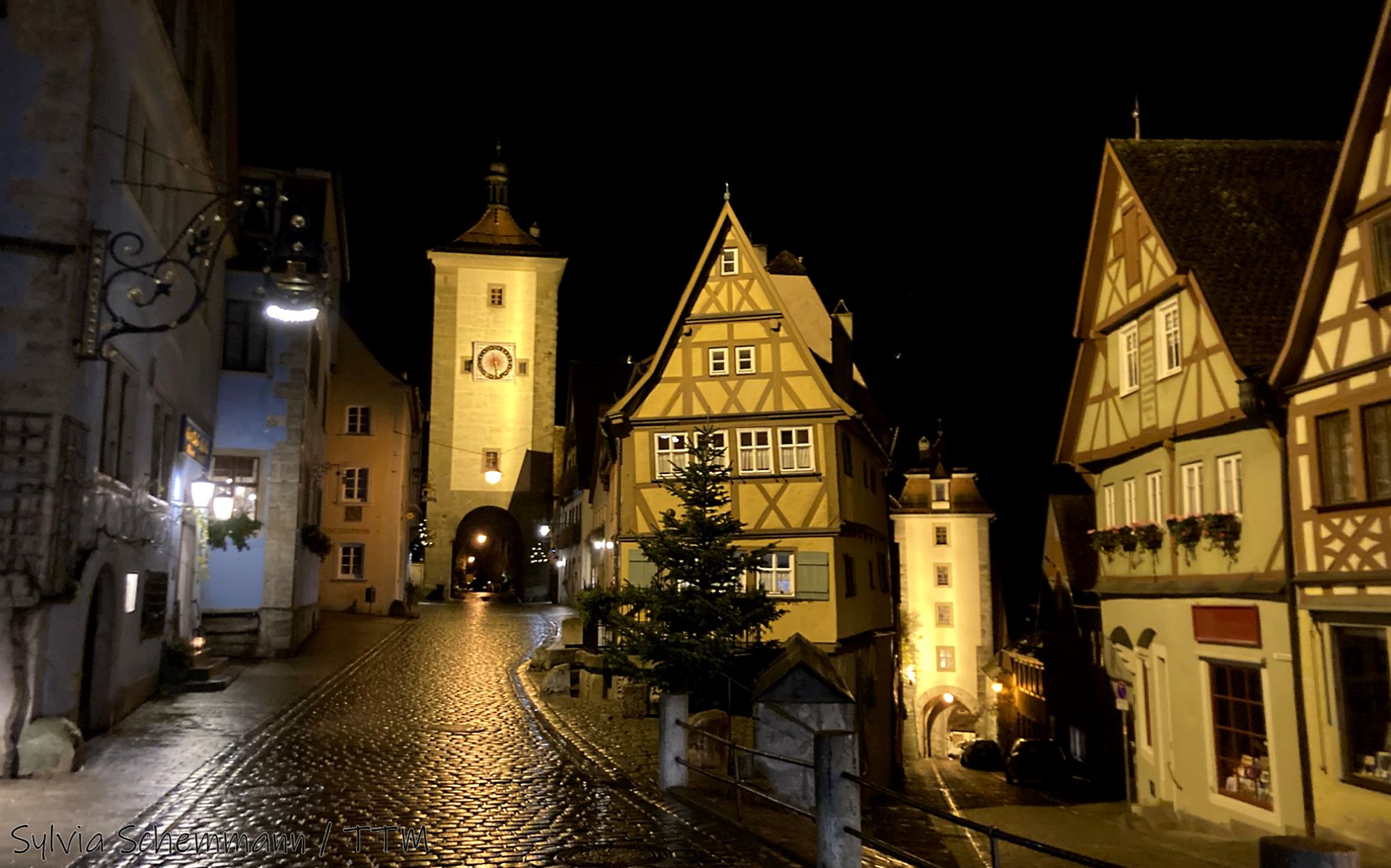 Blick auf einen historischen Turm, das Plönlein, in Rothenburg rechts davon sind Fachwerkhäuser zu sehen, vor einem dunklen Nachthimmel.