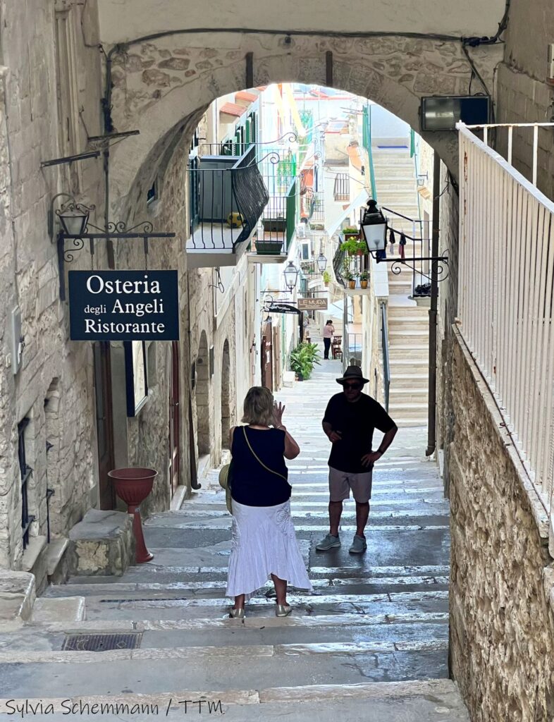 Eine Frau fotografiert einen Mann, der unter einem steinernen Bogen in der Altstadt von Vieste steht, hinter ihm ist eine Gasse zu sehen.