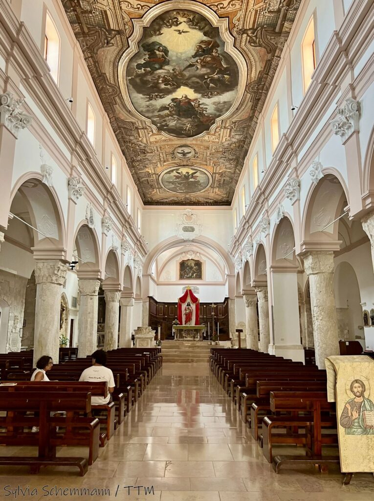 Blick in das Innere der Kathedrale von Vieste: Weiße Säulen, eine mit Gemälde verzierte Decke sowie braune Kirchenbänke aus Holz.