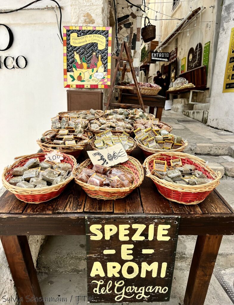 Ein Holztisch, auf dem mehrere Körbe stehen, in denen Tütchen mit Gewürzen und Kräutern sind, davor ein handbemaltes Schild mit der Aufschrift "Spezie e Aromi del Gargano"