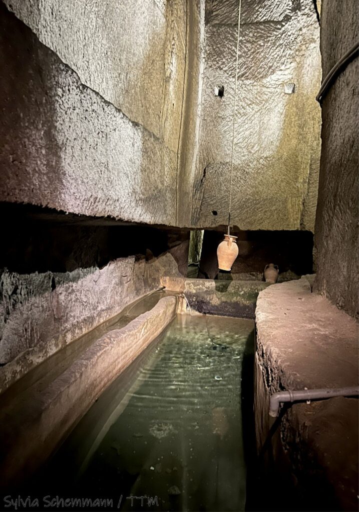 Ein schmales Wasserbecken, darüber hängt eine Amphore an einem Seil