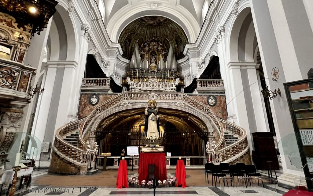 Eine doppelte geschwungene Treppe aus einem rötlichen Stein, die zur Orgel hochführt. Davor steht eine Heiligenfigur.